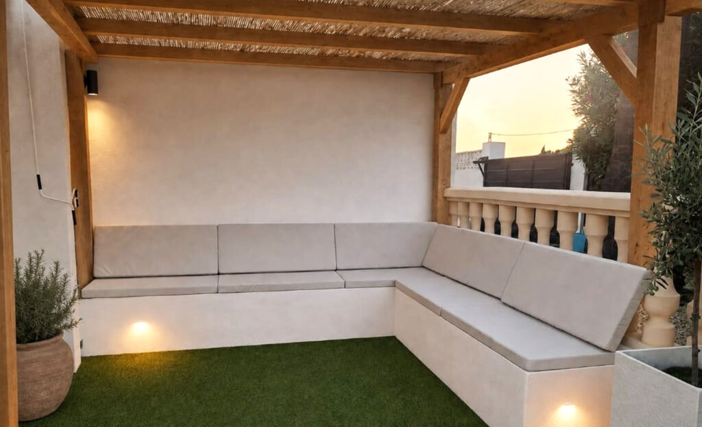 Rincón con espuma a medida en un banco en L, con asiento y respaldos tapizados en tono arena bajo una pérgola de madera